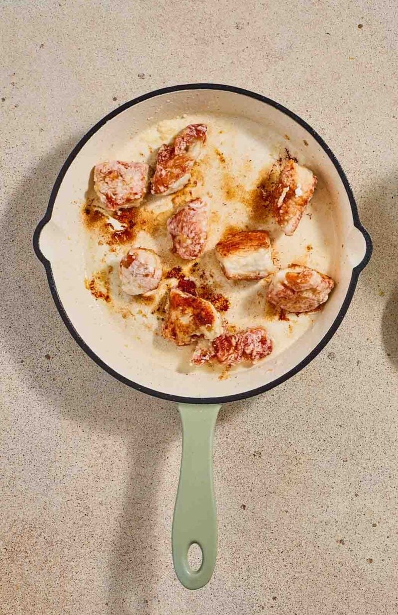 A skillet with browned chicken pieces sits below six small bowls containing green onions, sesame seeds, liquid, and powders on a beige surface.