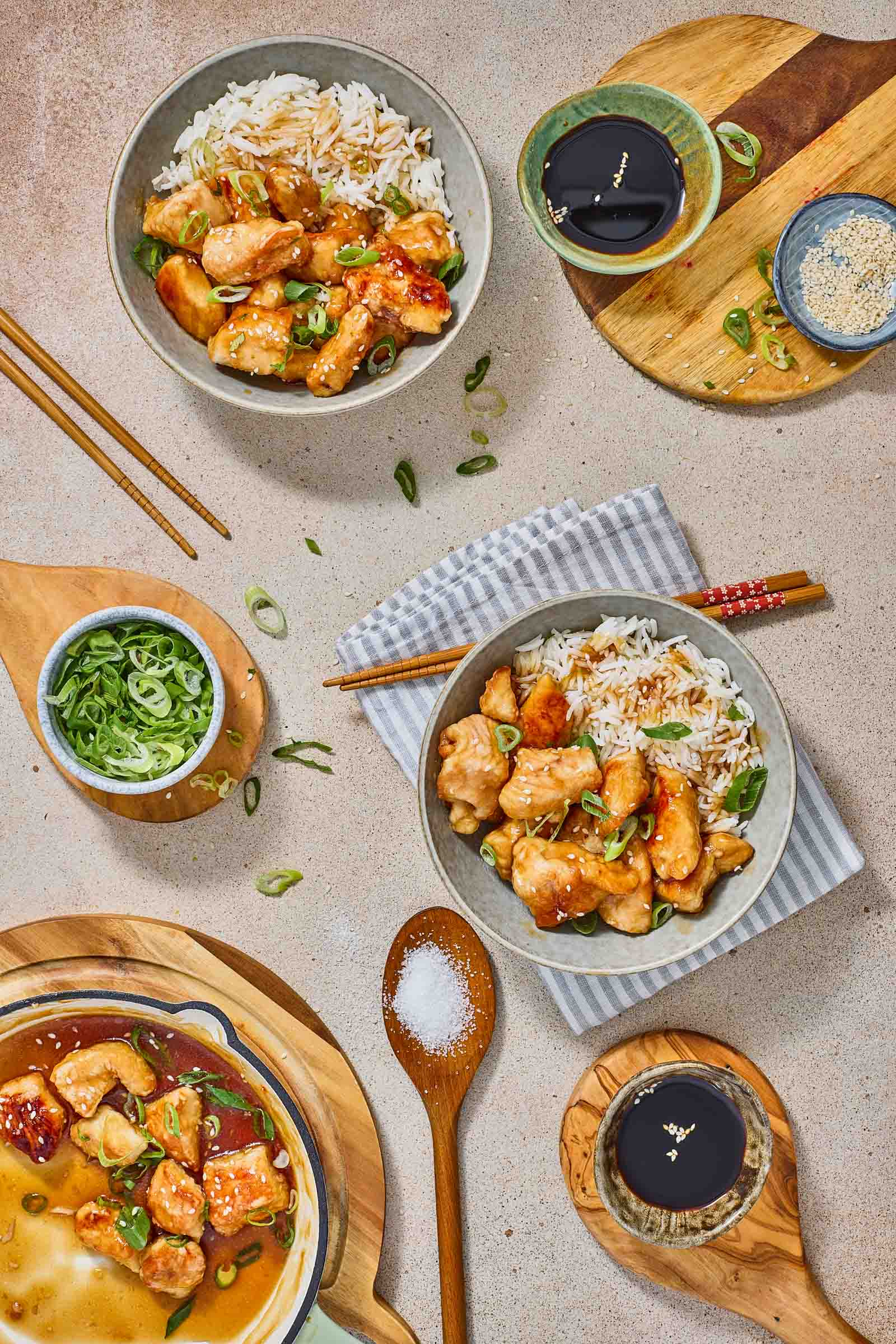 Three bowls of rice topped with sweet and sour chicken and sliced green onions are arranged on a table with chopsticks, sauce dishes, and a bowl of sesame seeds.