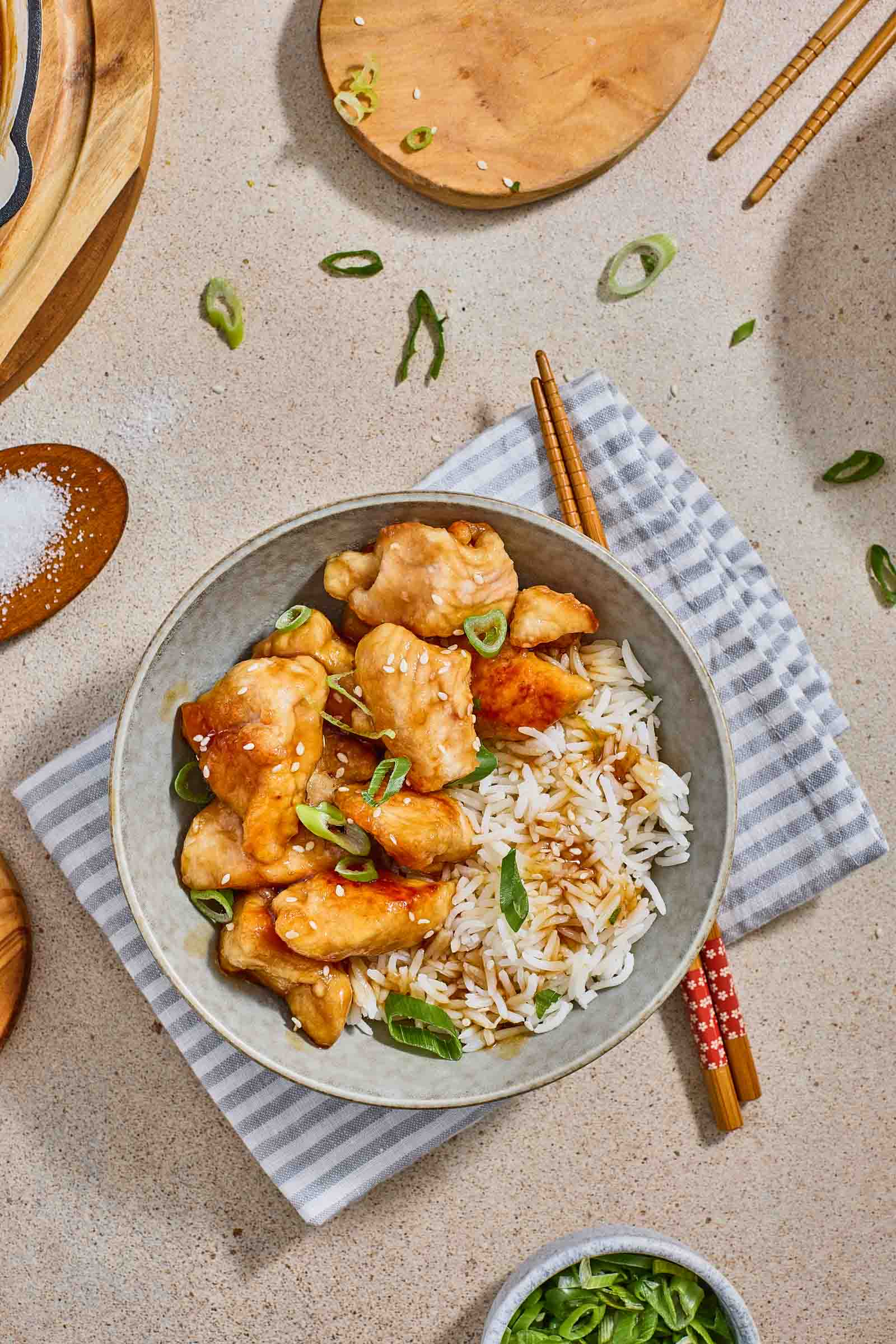 A bowl of white rice topped with glazed sweet and sour chicken pieces and garnished with chopped green onions, placed on a striped napkin with chopsticks beside it.