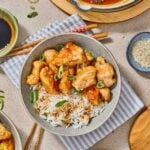 A bowl of sweet and sour chicken pieces over rice and sliced green onions, surrounded by dishes of sauce, sesame seeds, and more chicken on a set table.