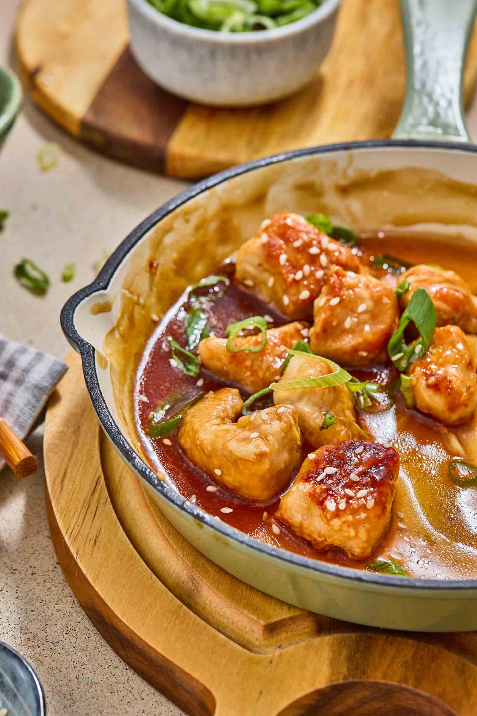 A skillet filled with glazed sweet and sour chicken pieces garnished with sesame seeds and sliced green onions, sitting on a wooden board.