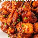 Close-up of sweet chili chicken bites garnished with chopped green onions, served on a white plate with a pair of chopsticks picking up a piece.