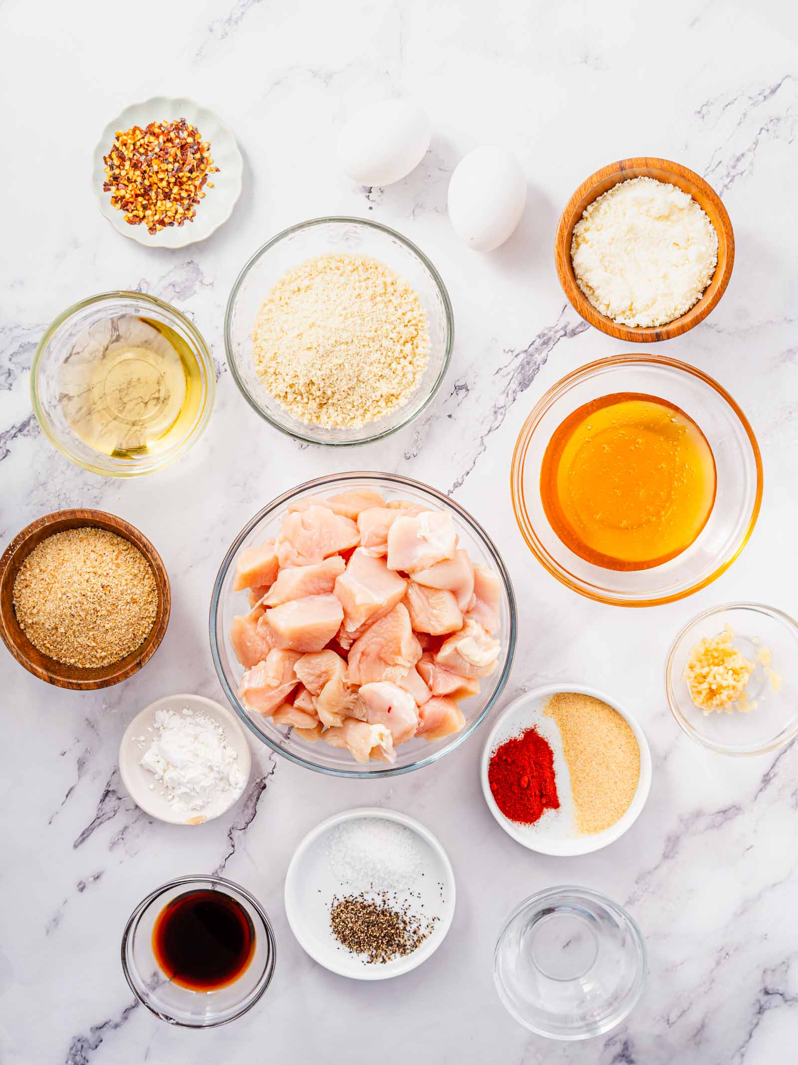 Overhead view of various ingredients in small bowls, including diced chicken, breadcrumbs, eggs, honey, spices, and sauces arranged on a white marble surface.