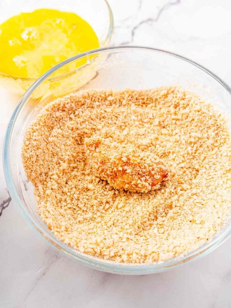 A piece of food being coated in breadcrumbs in a glass bowl, with a bowl of beaten egg visible in the background on a marble surface.