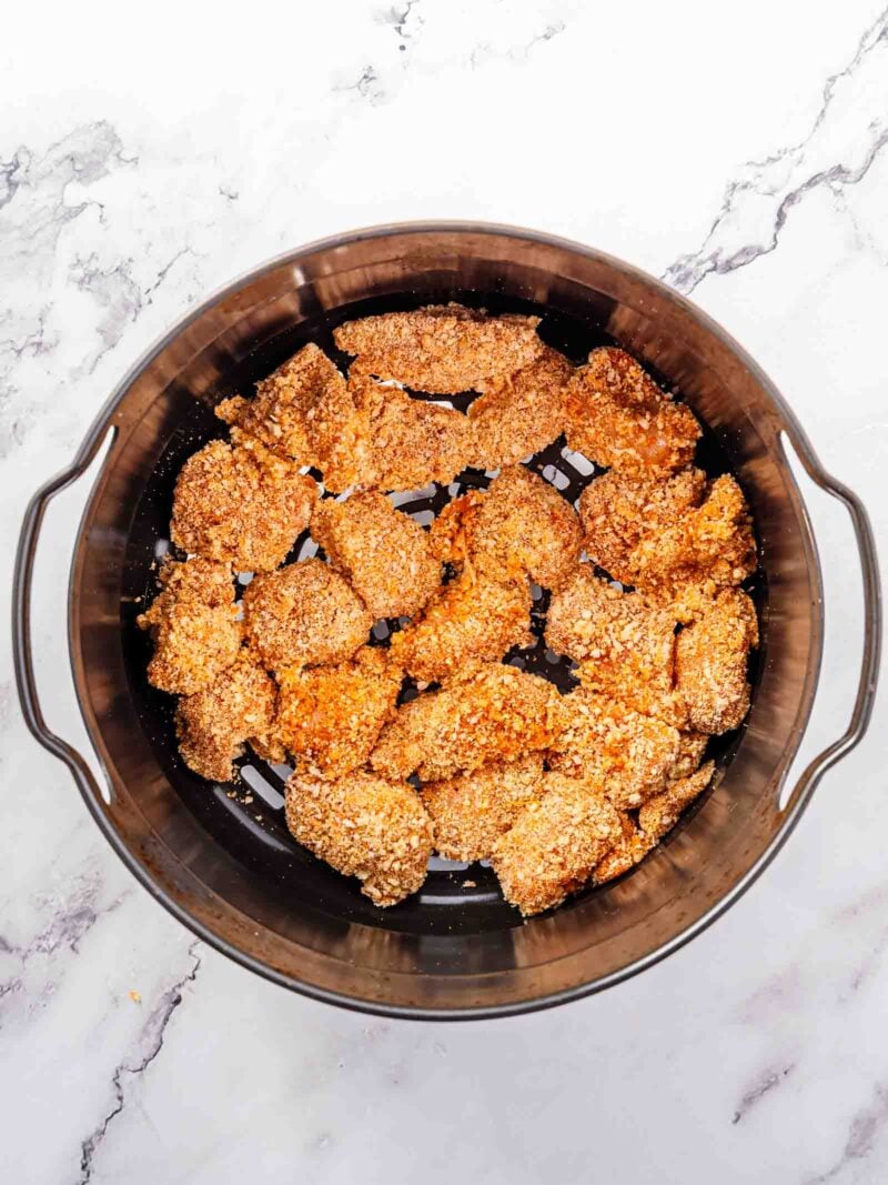Breaded chicken pieces arranged in a single layer inside an air fryer basket on a marble surface.