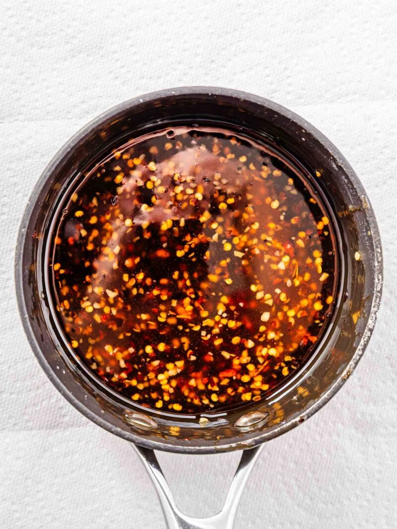 A saucepan filled with a dark sweet chili sauce containing visible red chili flakes, placed on a white textured surface.