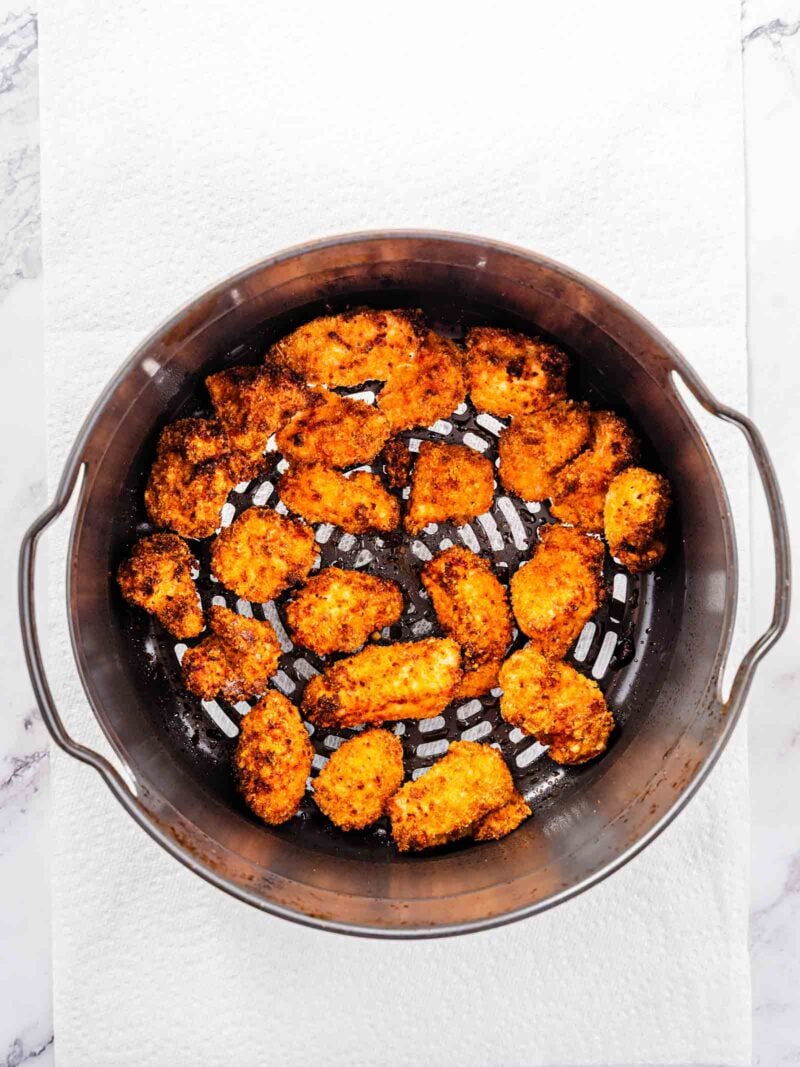 Round metal air fryer basket containing several pieces of breaded, cooked chicken resting on a white paper towel.