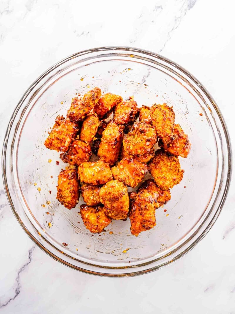 A clear glass bowl filled with breaded, seasoned, and chicken bites being sauced on a white marble surface.