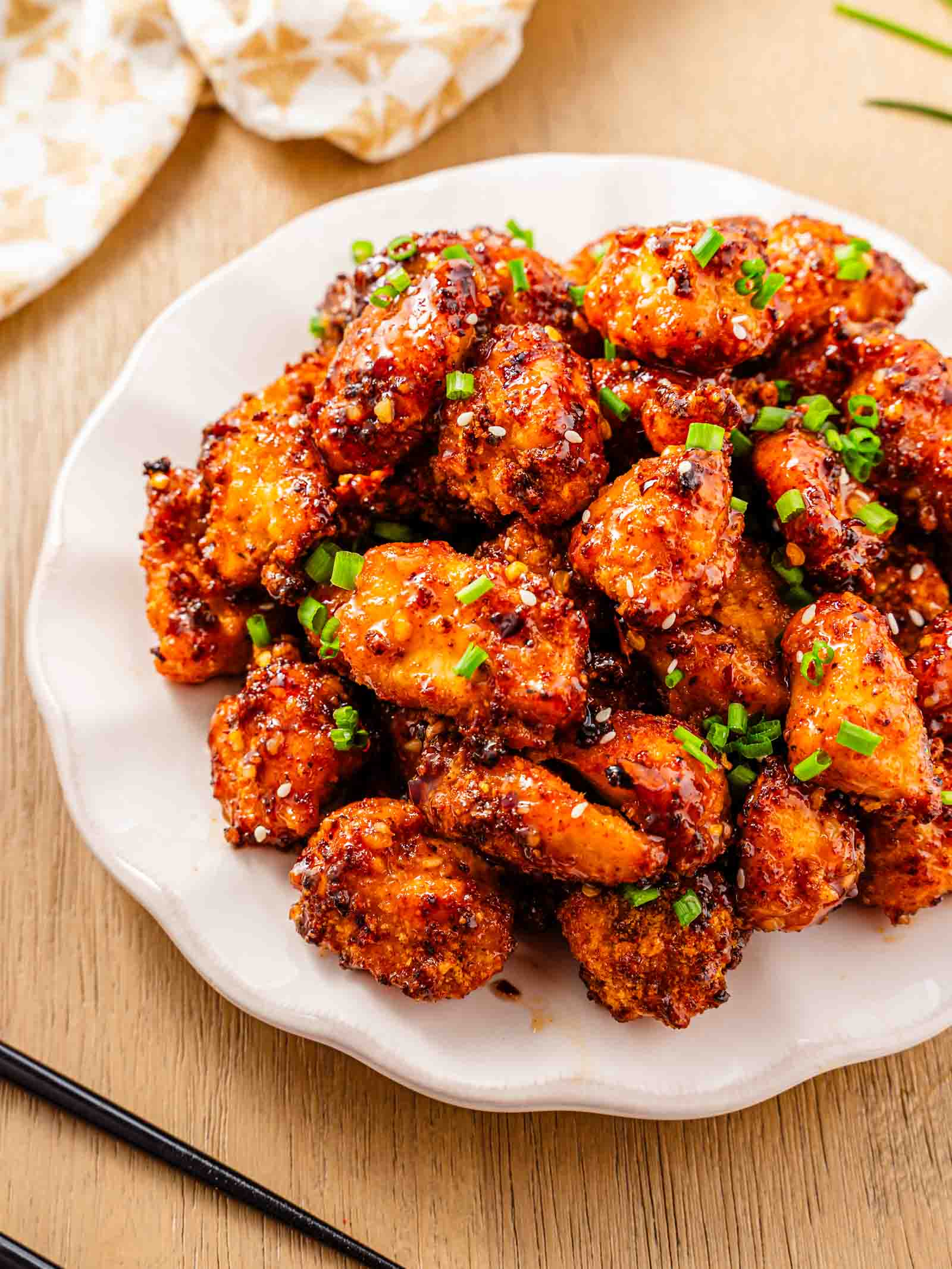 A white plate filled with crispy sweet chili chicken bites garnished with chopped green onions, placed on a wooden table with chopsticks and a cloth napkin nearby.