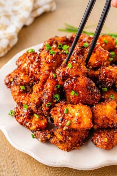 A plate of crispy, glazed sweet chili chicken bites garnished with chopped green onions, with chopsticks picking up one piece.