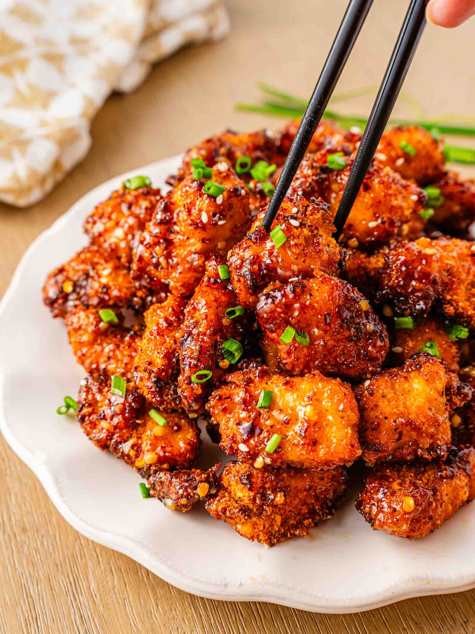 A plate of crispy, glazed sweet chili chicken bites garnished with chopped green onions, with chopsticks picking up one piece.