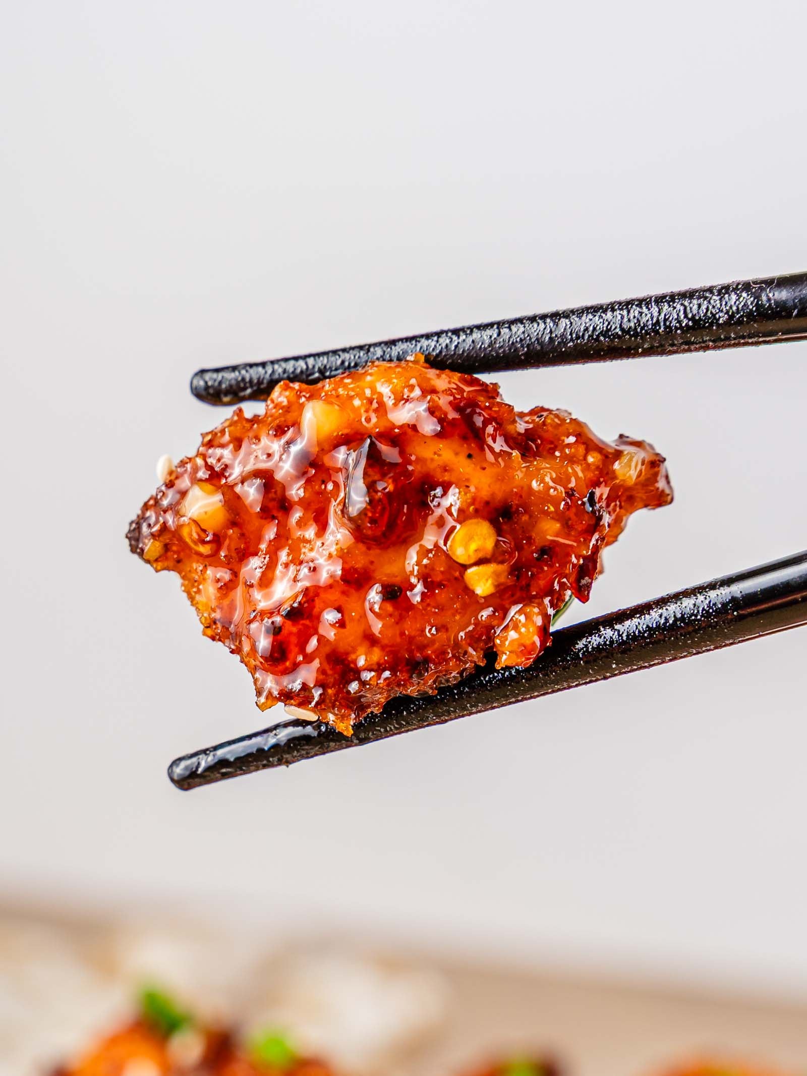 A close-up of chopsticks holding a glazed, sweet chili chicken bite with visible chili flakes against a plain background.