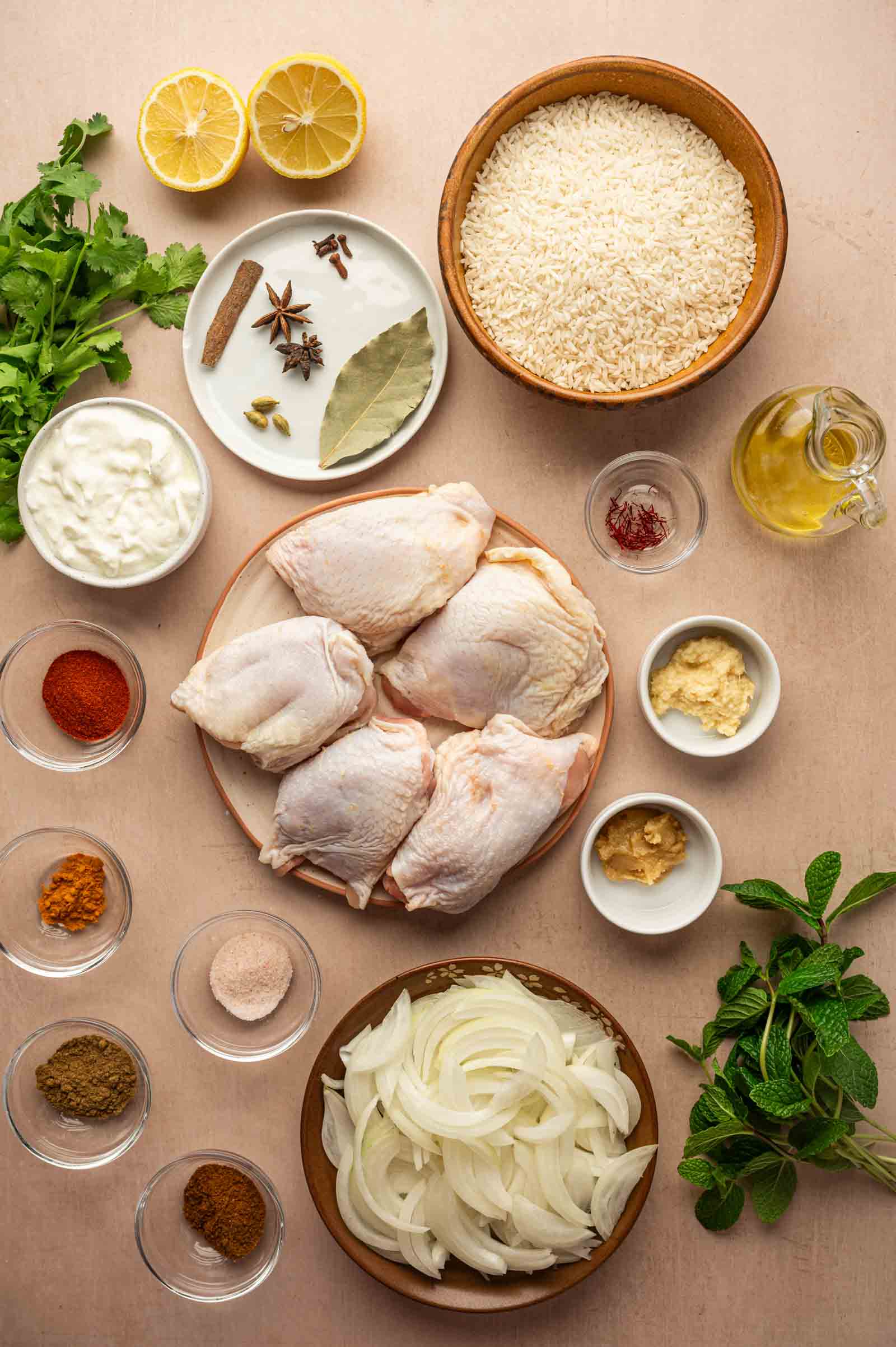 Overhead view of raw chicken thighs, bowls of rice, sliced onions, yogurt, spices, oil, garlic, ginger, fresh cilantro, mint, and lemon halves arranged on a light surface.