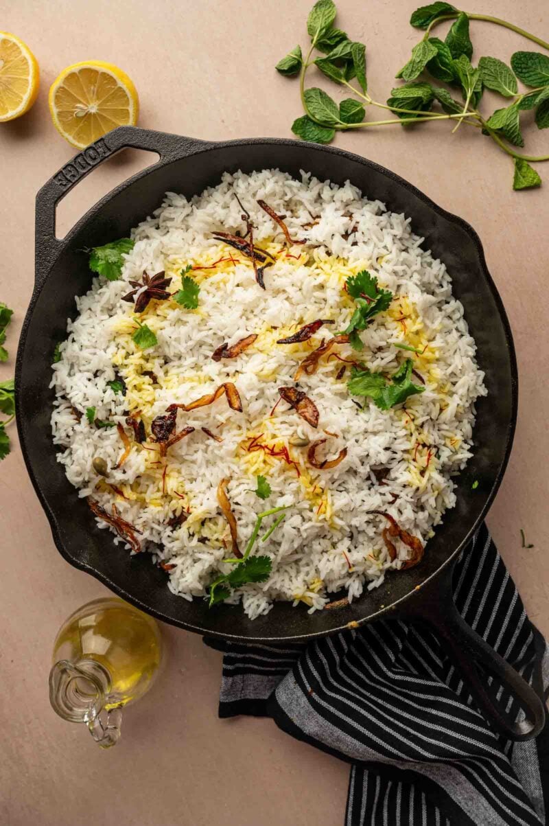 A cast iron pan filled with cooked basmati rice, garnished with fried onions and fresh cilantro, sits on a striped cloth with lemon halves, mint leaves, and a bottle of oil nearby.