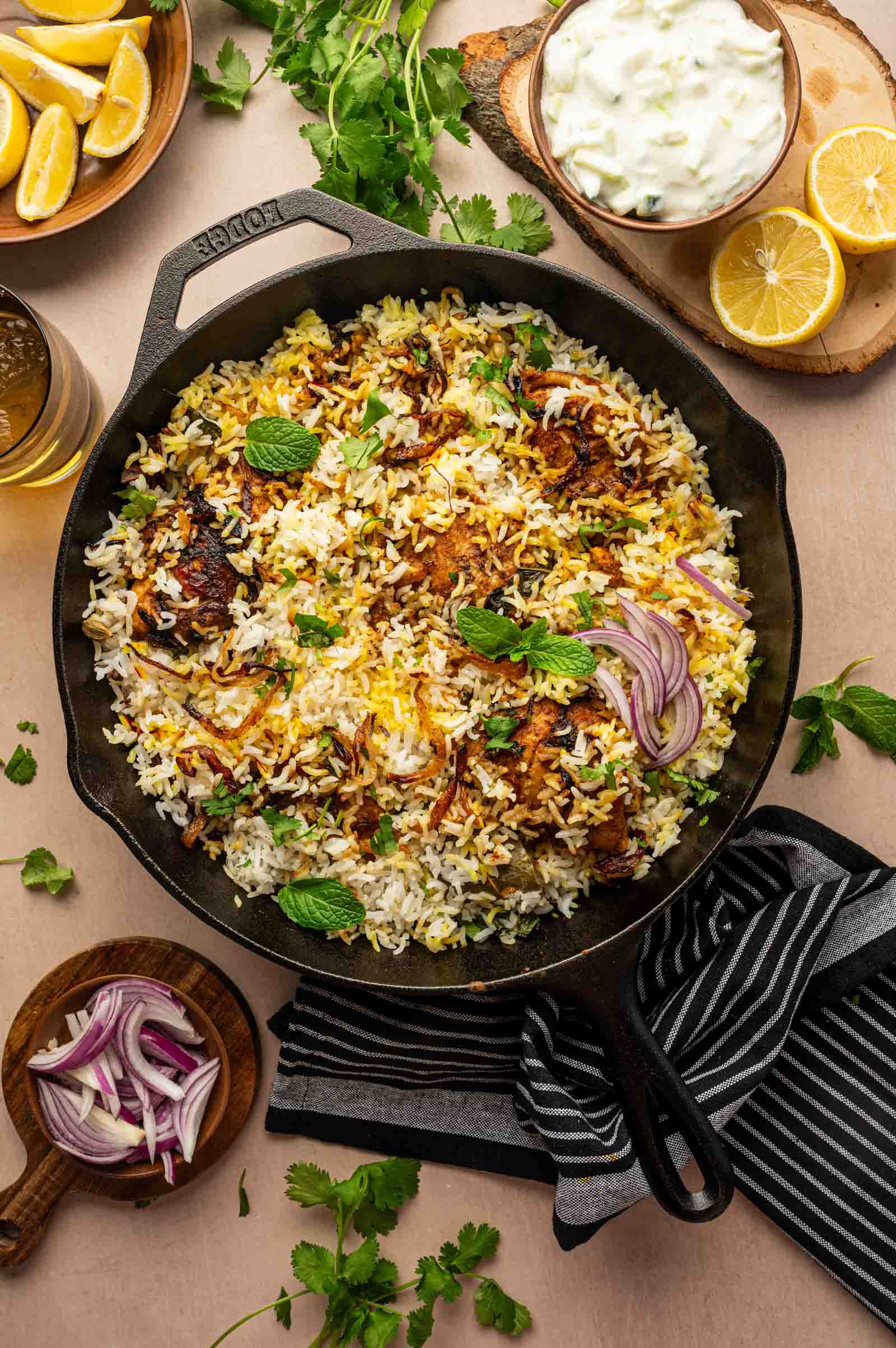 A cast iron skillet filled with chicken biryani garnished with mint leaves and sliced red onions, surrounded by lemon wedges, cilantro, and a bowl of yogurt sauce.