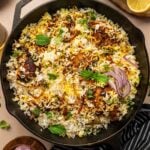 A cast iron skillet filled with chicken biryani, garnished with fresh herbs and sliced red onions, sits on a table with lemon halves, yogurt sauce, and herbs nearby.