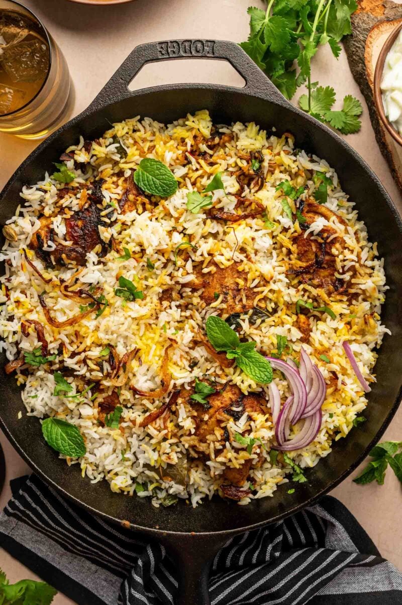 A cast iron skillet filled with chicken biryani, garnished with fresh mint leaves, cilantro, and sliced red onions.