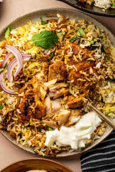 A plate of chicken biryani with sliced red onions, fresh mint leaves, and a dollop of yogurt on the side.