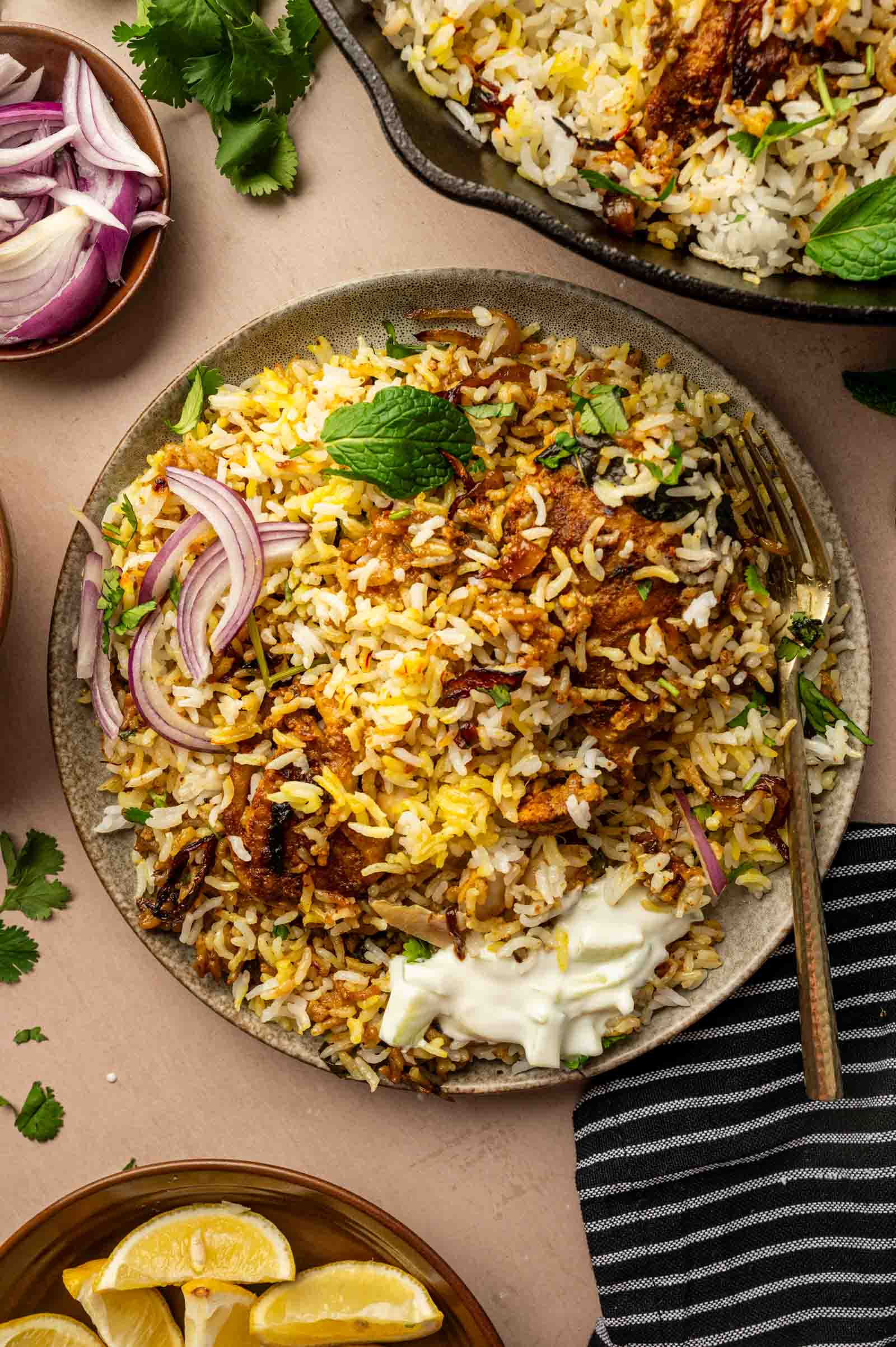 A plate of chicken biryani rice with pieces of meat, garnished with red onion slices, fresh herbs, a dollop of yogurt, and served with lemon wedges and extra onions on the side.