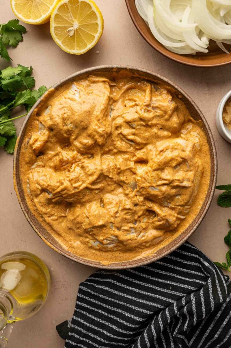 A bowl of marinated chicken in a creamy orange sauce, surrounded by sliced onions, lemon halves, cilantro, olive oil, and herbs on a light surface.
