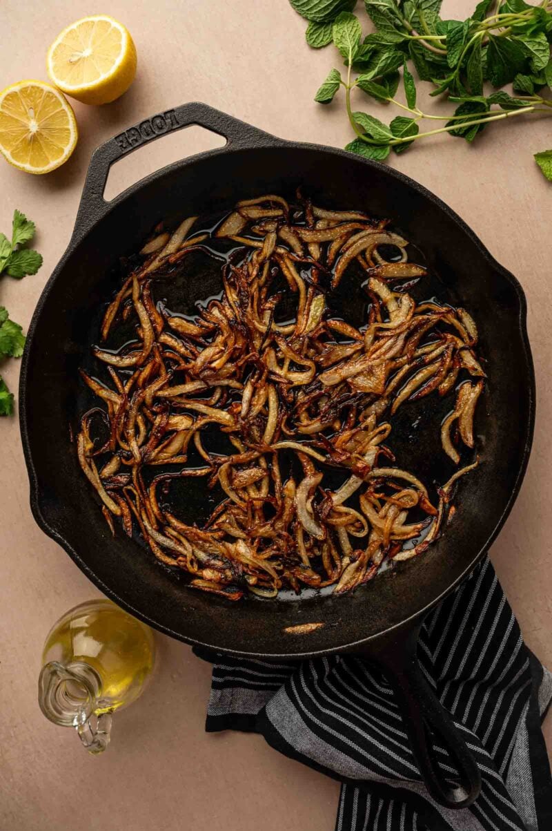 A cast iron skillet with caramelized onions, surrounded by a halved lemon, fresh mint, and a bottle of olive oil on a light surface.