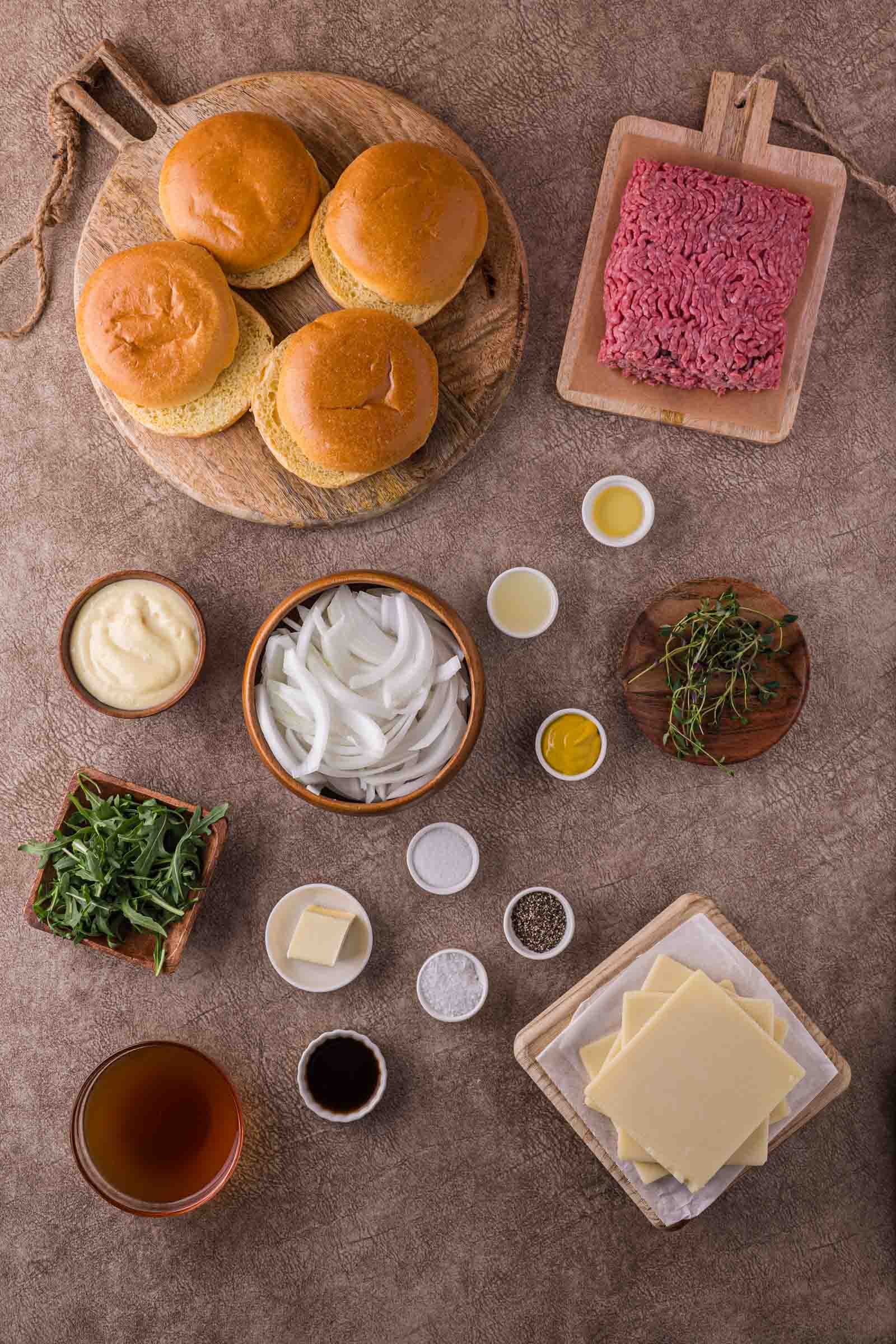 Top-down view of burger ingredients including buns, ground beef, cheese slices, sliced onions, arugula, thyme, butter, condiments, seasonings, and a beverage on a brown surface.