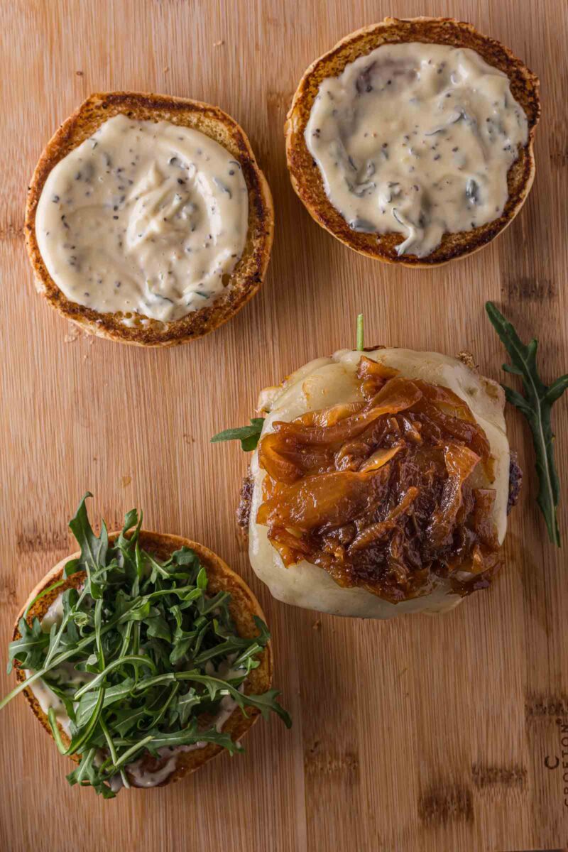 Open burger on a wooden board with toasted buns, mayonnaise, arugula, melted cheese, and caramelized onions on a beef patty.
