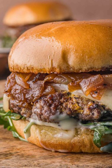 Close-up of a cheeseburger with caramelized onions, melted cheese, lettuce, and sauce in a glossy toasted bun, sitting on a wooden surface.