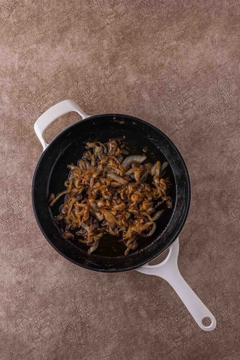 Sautéed caramelized onions in a black pan with a white handle, placed on a textured brown surface.