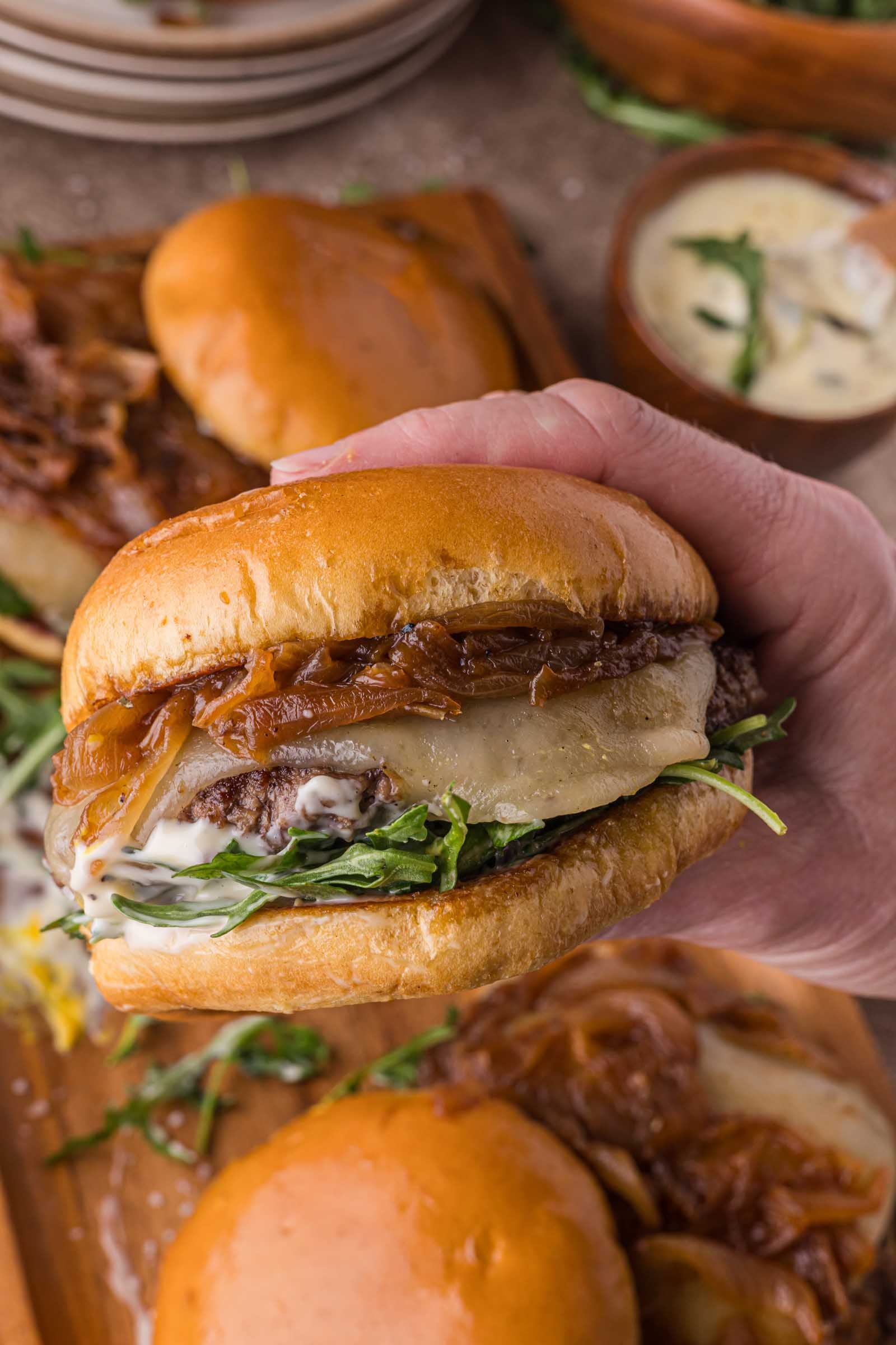 A hand holds a burger with caramelized onions, melted cheese, arugula, and sauce in a shiny bun. Other burgers and a bowl of sauce are visible in the background.