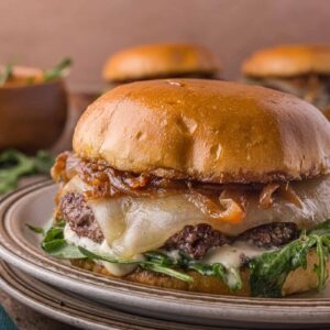 A cheeseburger with melted cheese, caramelized onions, greens, and sauce on a toasted bun sits on a plate.
