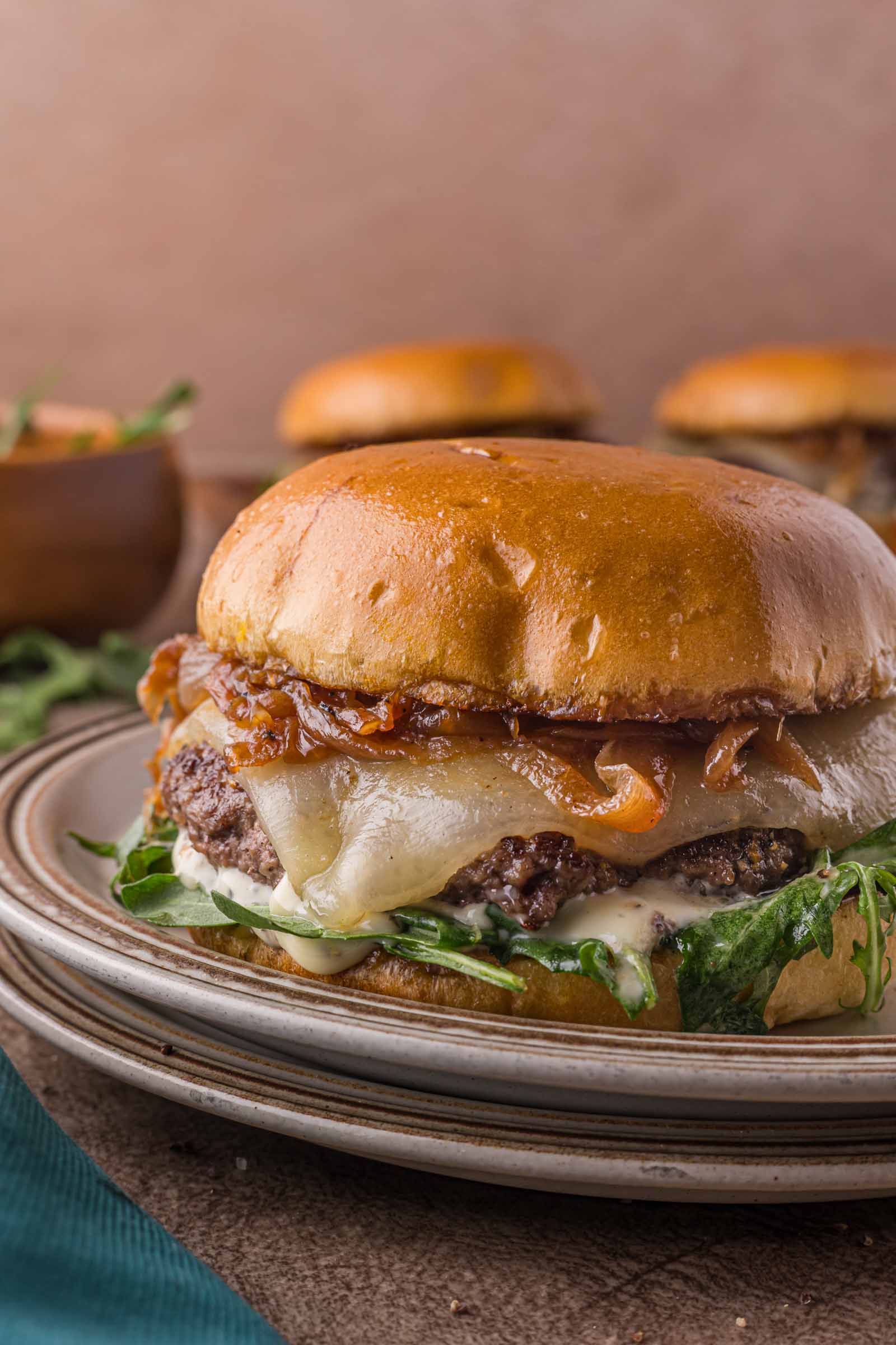A cheeseburger with melted cheese, caramelized onions, greens, and sauce on a toasted bun sits on a plate.