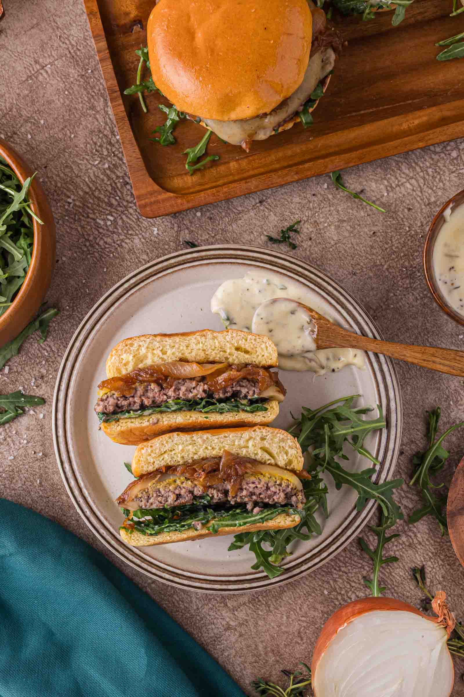 A sliced burger with arugula and onions on a plate, served with creamy aioli; another whole burger and bowls of arugula and sauce are nearby.