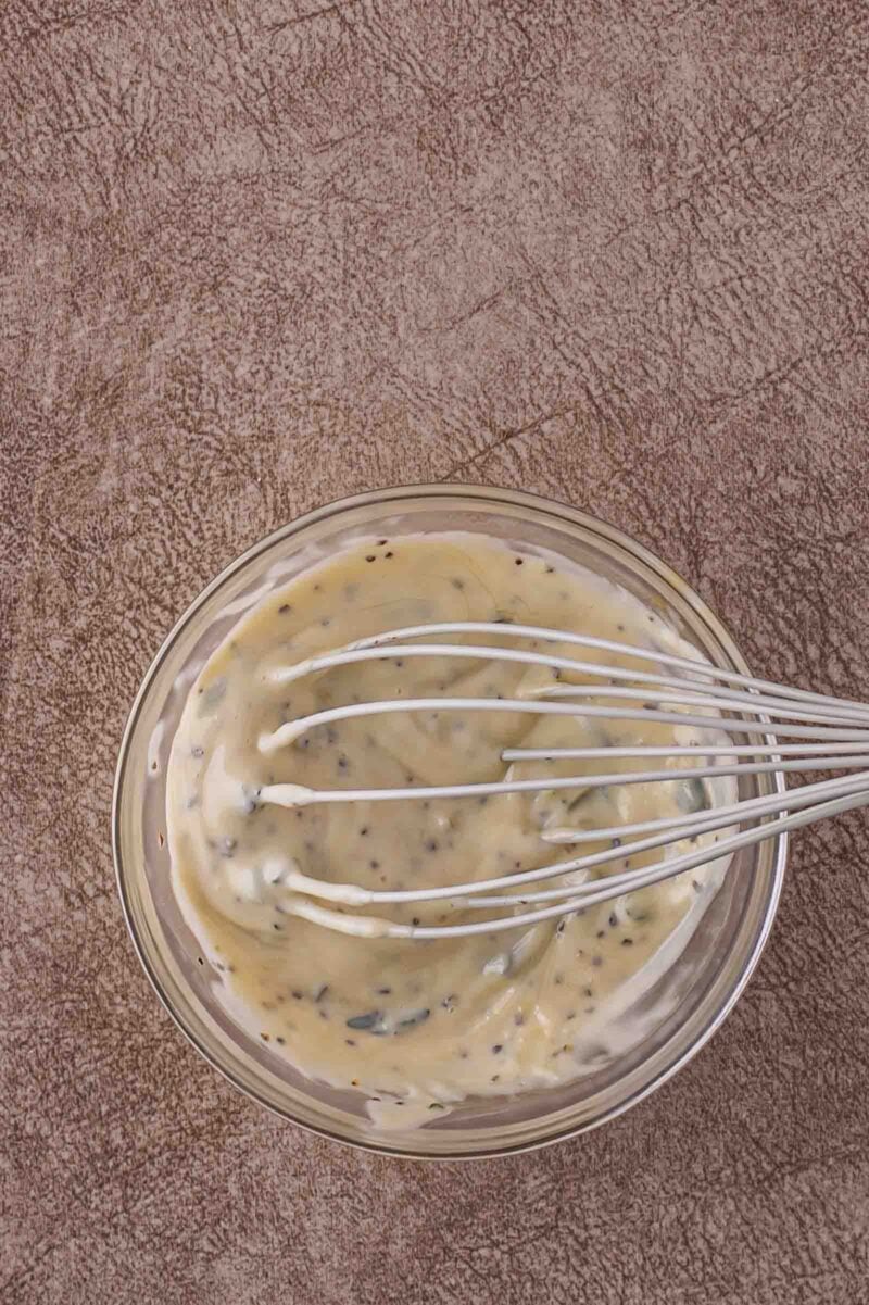 A metal whisk rests in a glass bowl filled with a creamy, thyme aioli sauce on a textured brown surface.