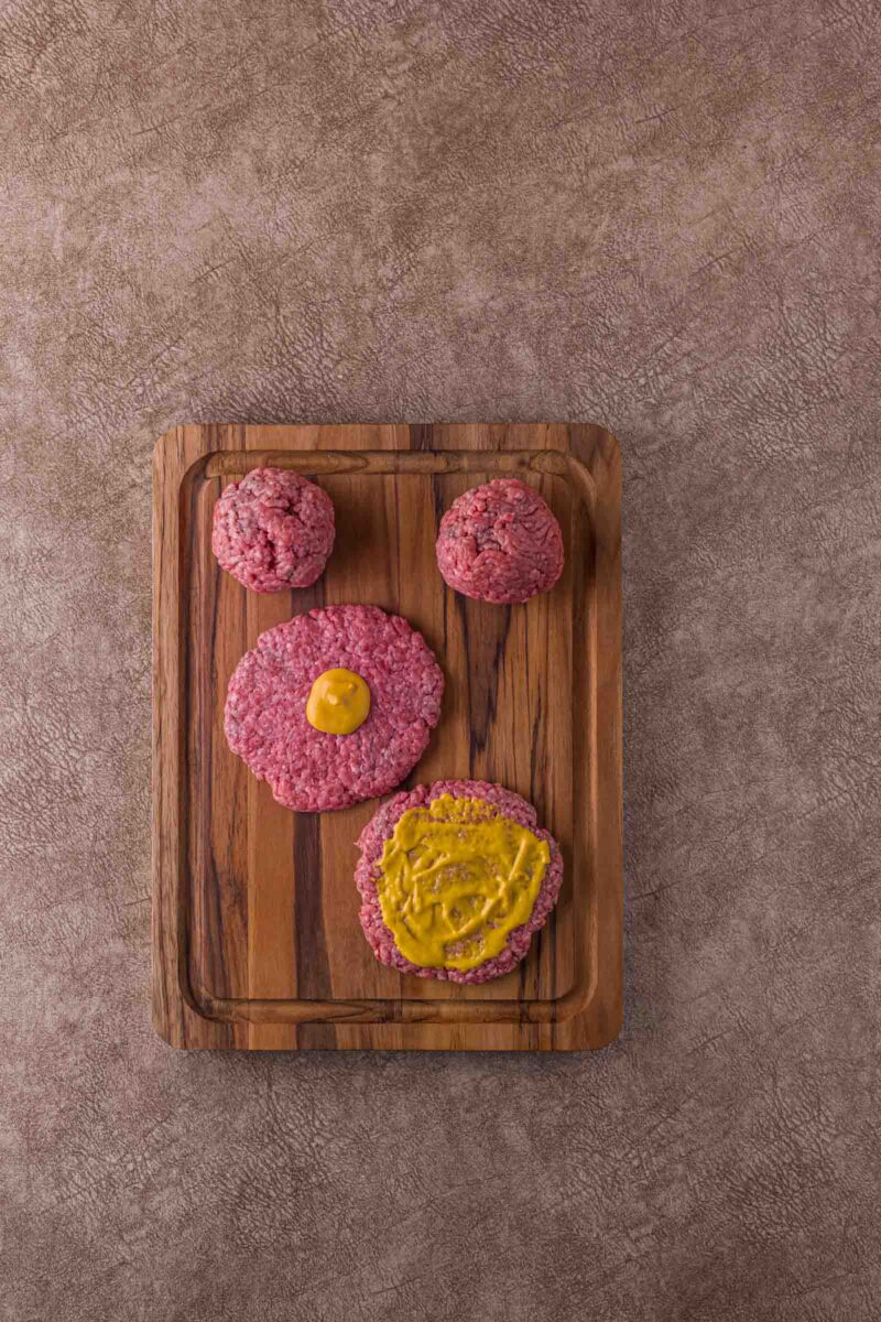 Two raw hamburger patties and two raw meatballs on a wooden board, one patty topped with a thin layer mustard and another with a small amount of mustard.