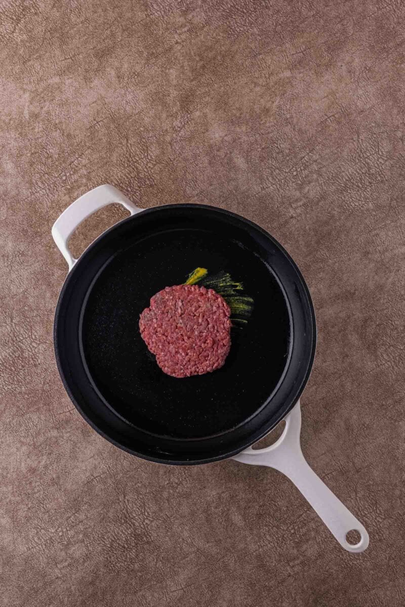 Raw hamburger patty placed in the center of a black frying pan with a white handle, on a textured brown surface.