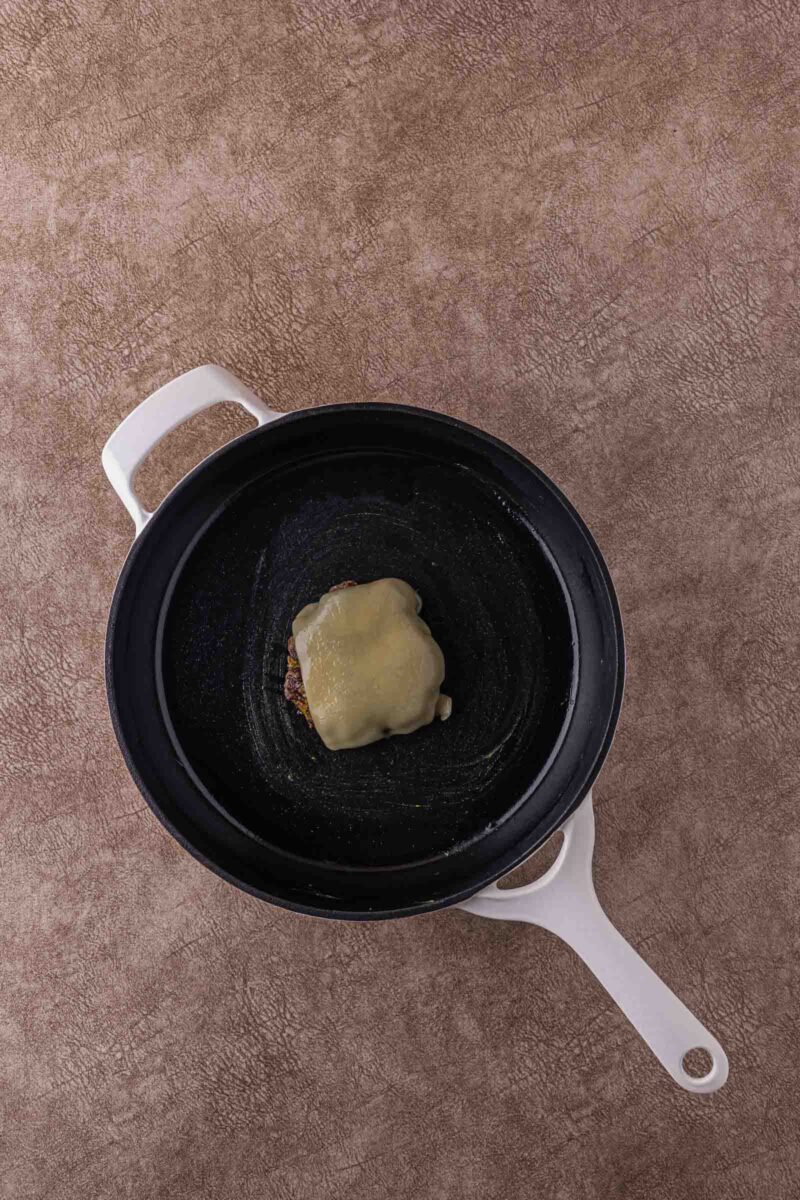 A single hamburger patty topped with melted cheese sits in the center of a black skillet with a white handle on a textured brown surface.