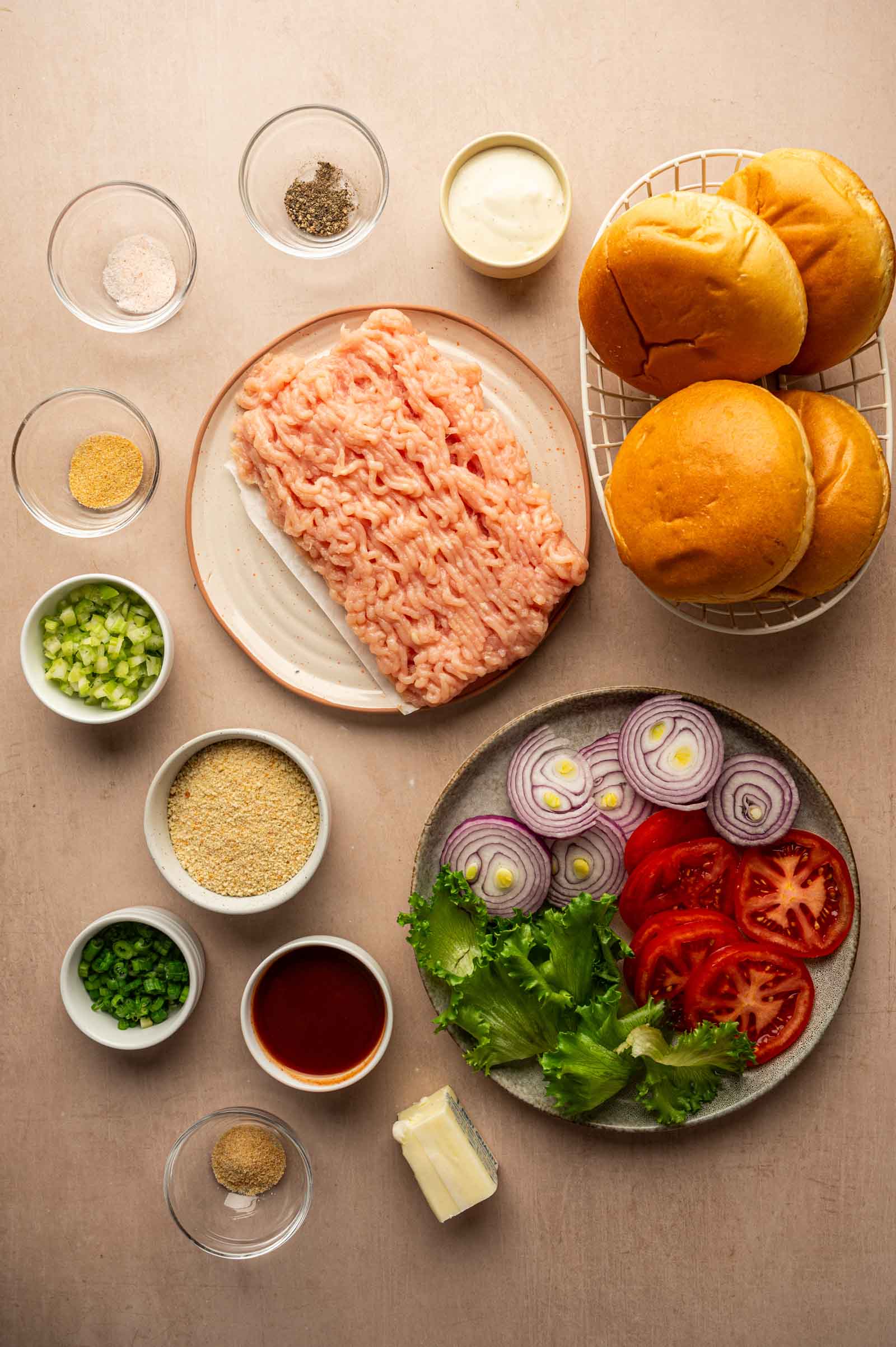 Ingredients for buffalo chicken burgers arranged on a table, including ground chicken, buns, sliced tomato, lettuce, red onion, seasonings, breadcrumbs, green onions, celery, and butter.