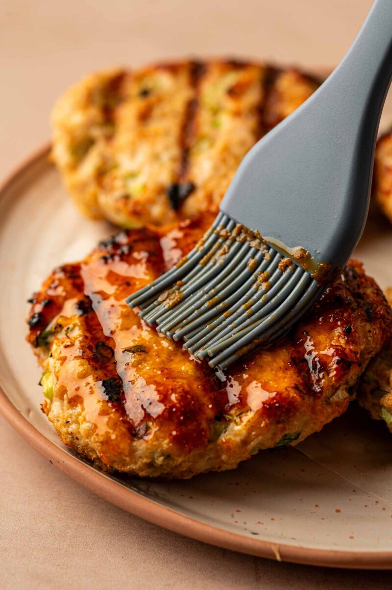 A silicone brush coats a grilled chicken burger with buffalo sauce on a ceramic plate.
