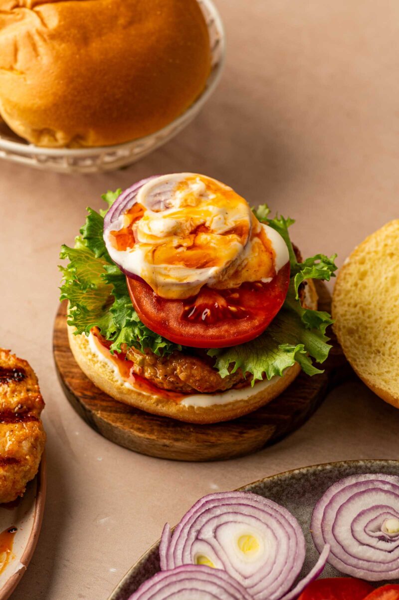 An open-faced grilled chicken burger with lettuce, tomato, onion, ranch dressing, and buffalo sauce on a bun, next to sliced onions and a plate of fried chicken.