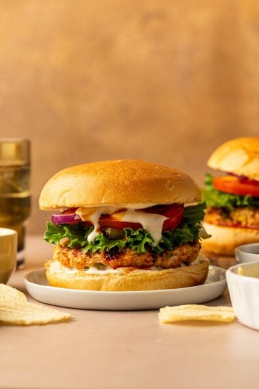 A buffalo chicken burger with lettuce, tomato, red onion, and sauce on a bun is served on a plate with potato chips, with another burger and drinks in the background.