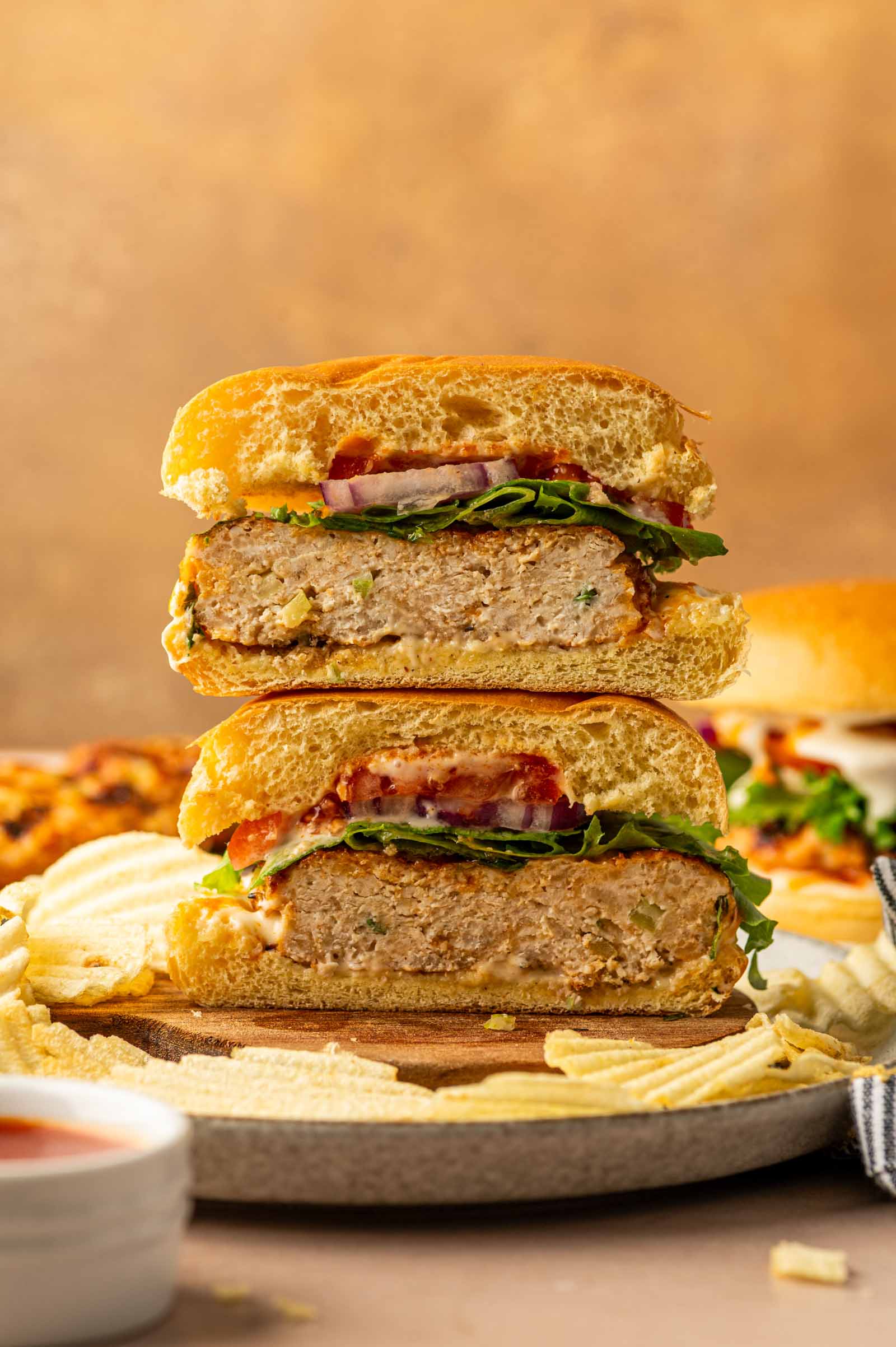 A grilled buffalo chicken burger cut in half and stacked, showing the inside with lettuce, tomato, onion, and sauce, served on a plate with potato chips.