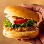 A hand holding a grilled buffalo chicken burger with lettuce, tomato, onions, and ranch in a bun against a plain background.