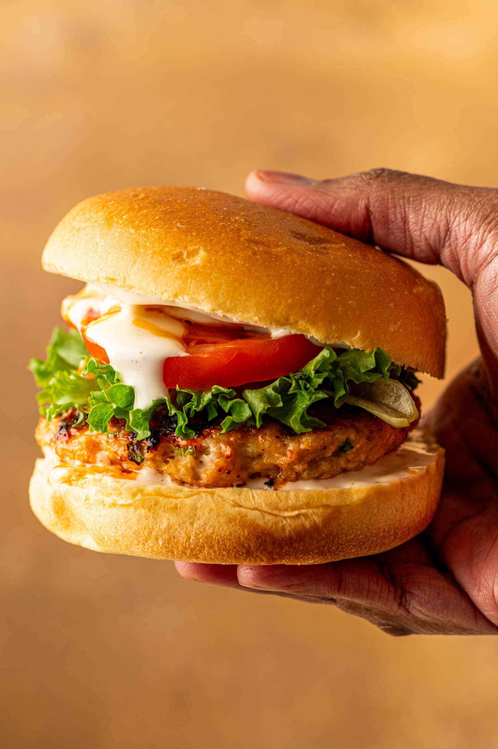 A hand holding a grilled buffalo chicken burger with lettuce, tomato, onions, and ranch in a bun against a plain background.