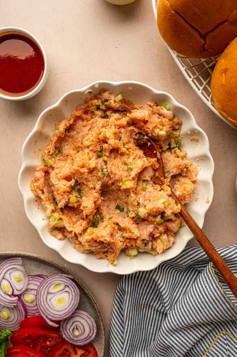 A bowl of raw ground chicken with diced green onions and celery, stirred with a wooden spoon, surrounded by burger buns, sliced onions, tomatoes, and a cup of sauce.