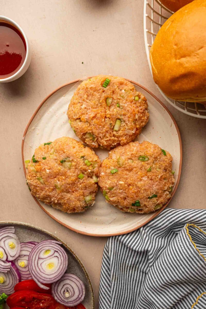 Three uncooked chicken burger patties with chopped green onions on a plate, surrounded by burger buns, sliced onions, tomatoes, and a bowl of sauce.