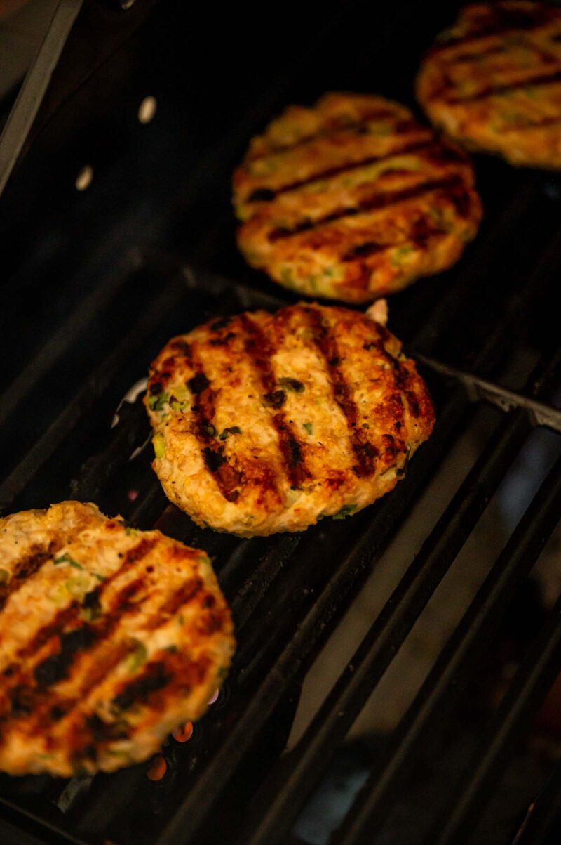 Four grilled chicken burger patties with visible grill marks are cooking on a barbecue grill.