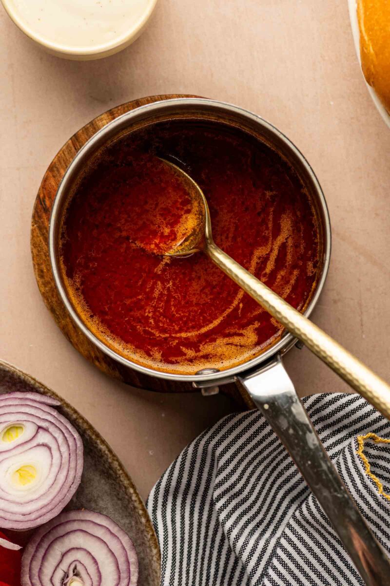 A saucepan filled with buffalo sauce and a spoon rests on a wooden coaster, next to sliced red onions and a striped cloth.