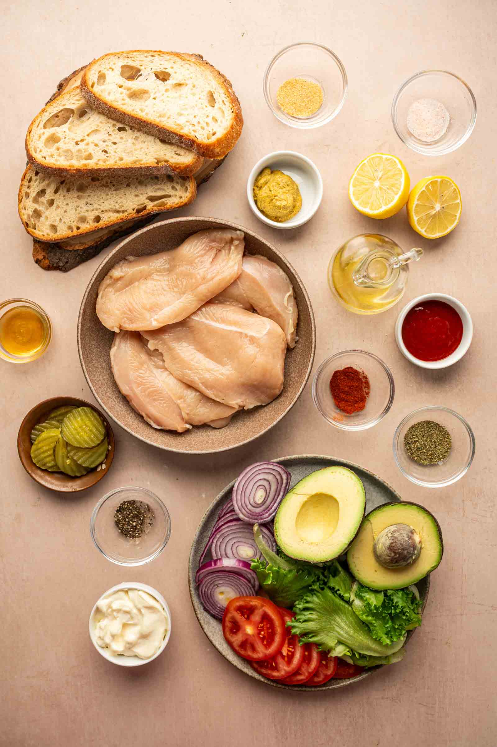 Sliced bread, raw chicken breasts, avocado, tomato, red onion, lettuce, pickles, mayonnaise, spices, mustard, lemon, oil, and sauce arranged on a beige surface.