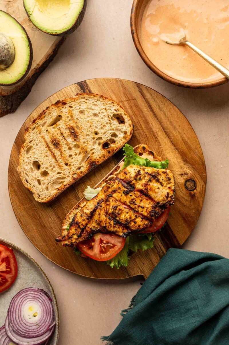 Grilled chicken breast on a slice of bread with lettuce and tomato, next to another bread slice, with avocado, onions, tomatoes, and sauce on the side.